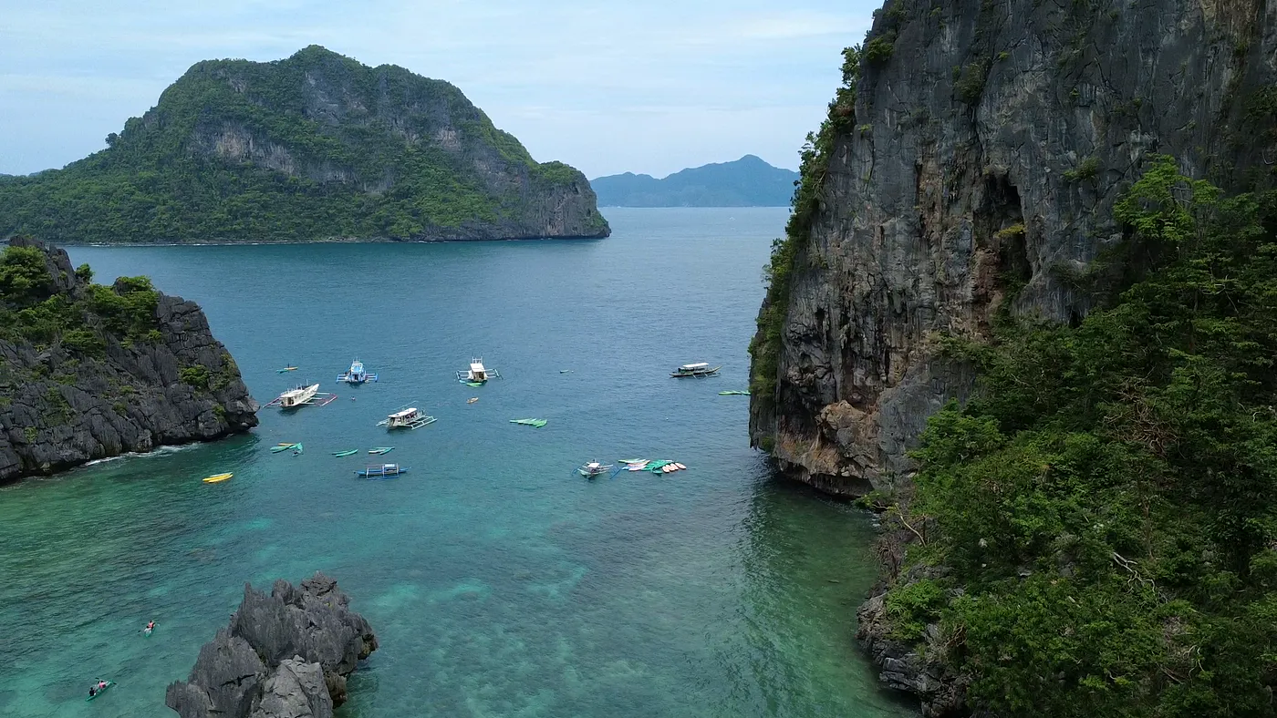 Aerial view of a channel between limestone cliffs opening onto turquoise water with outrigger boats, Palawan
