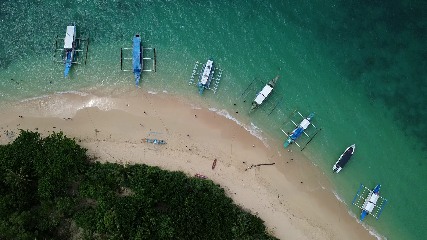 Top-down aerial of traditional outrigger boats moored along a curved white sand beach, Palawan