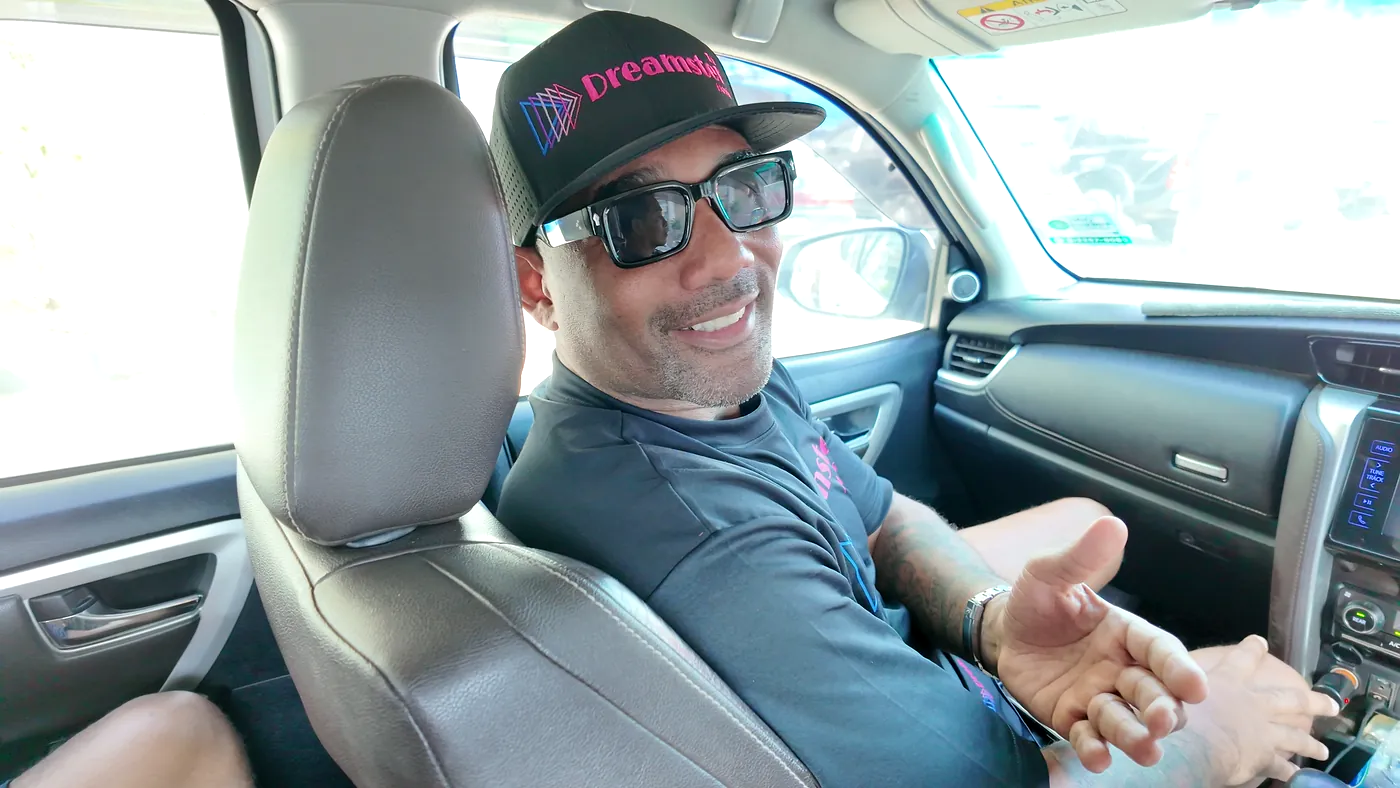 A man in a cap and sunglasses smiling and gesturing in the front seat of a car, filmed from the back seat