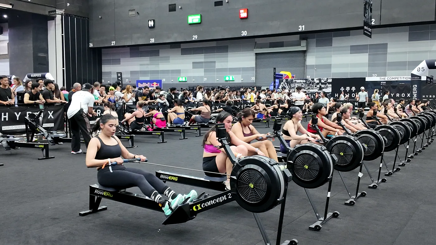 Rows of Concept2 RowErg machines on the HYROX Bangkok competition floor, athletes mid-race with spectators lining the barrier behind